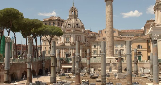 A closeup view of the Trajan's Forum archaeological site in Rome, Italy, displaying weathered granite columns standing among ancient stone fragments, brick structures, and a historical church.