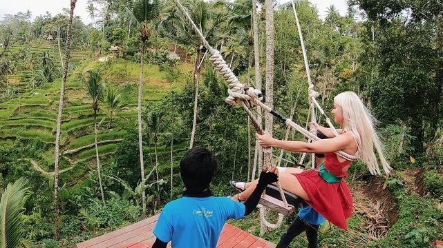 Rear view of blonde woman seated on rope swing above steep terraced valley, framed by tall palm trunks, dense jungle, green hillside, and two observers beside wooden platform in calm adventure mood