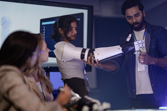 An Indian woman presents a robotic arm prototype to her diverse tech team. A Caucasian woman listens intently during the late-night innovation meeting in a modern office.