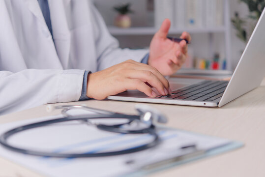 male doctor physician white coat typing laptop computer keyboard desk stethoscope clipboard foreground hospital clinic healthcare professional online consultation electronic medical record working