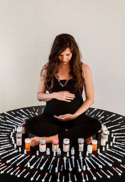 Pregnant woman sitting cross-legged surrounded by IVF syringes and medicine bottles.
