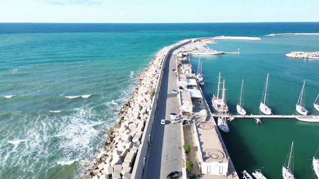 Vieste Marina, Gargano, Puglia, Italy. Aerial drone footage following a car along the harbor breakwater with sailboats, yachts and turquoise Adriatic Sea.