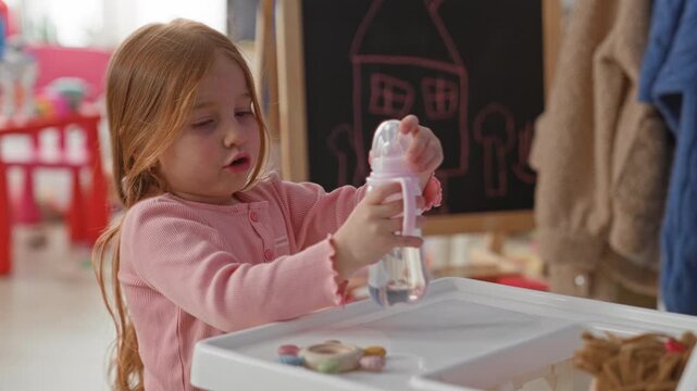 Girl holding baby bottle and examining small pastel toys on a plastic tray in building play area; playful curiosity.