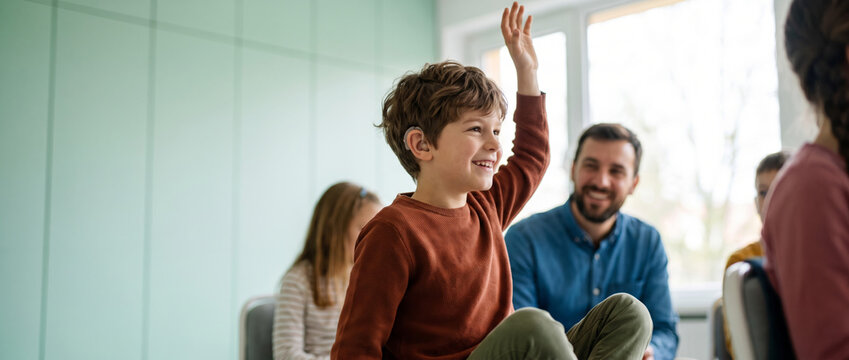 Boy with a hearing aid participating in a classroom discussion. Family support group or teacher visible in the background. Concept of pediatric hearing care and inclusive learning.