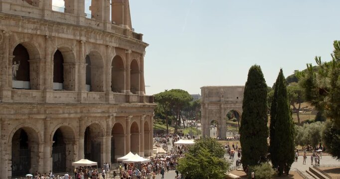 A stunning perspective next to the ancient Colosseum facade, looking towards the nearby triumphal Arch of Constantine surrounded by tall green cypress trees and bustling tourists in Rome, Italy.