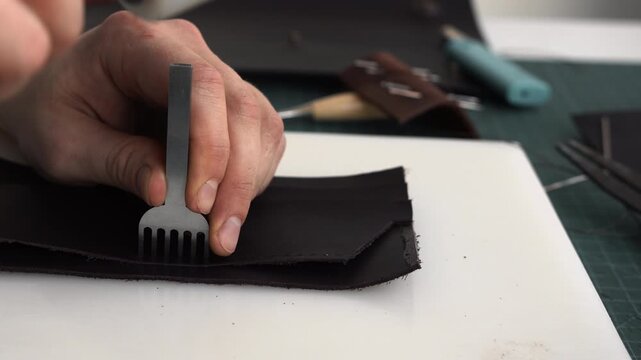 Craftsman perforating black leather with a stitching chisel and a maul in a workshop. Creating a handmade accessory