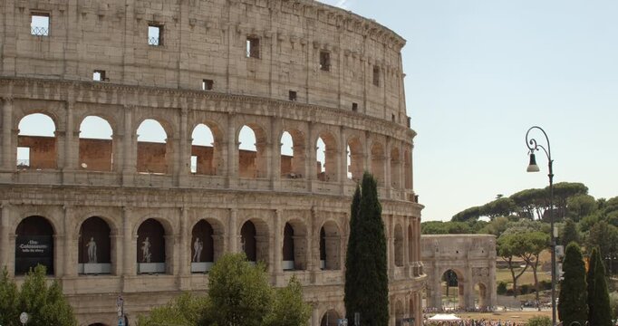 Panorama of the majestic Colosseum in Rome under a sunny sky, showing its classic stone arches alongside the Arch of Constantine, cypress trees, and a vintage street lamp post in Italy.