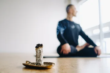 Fototapeten Lotusblume A male athlete in black sportswear sits in the lotus position against the background of an incense candle in the gym.  © Alex