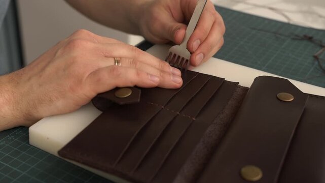 Craftsman punching stitching holes in a handmade brown leather wallet using a pricking iron tool
