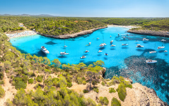 Aerial drone view of luxury yacht, boats in turquoise sea water bay of Mallorca, Spain in summer. Motorboats and sailboats in mediterranean sea cove near sand beach, green trees on rocky coast