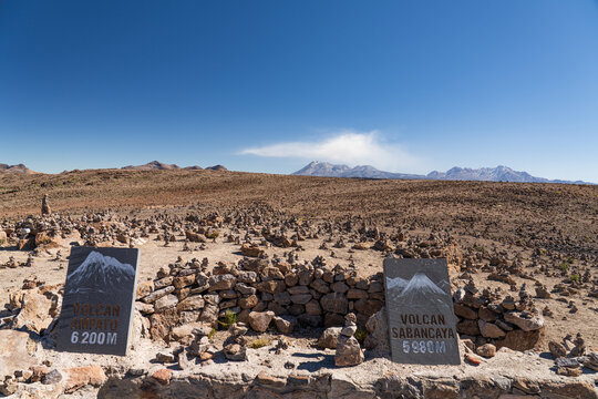 Stone signs for the Ampato and Sabancaya volcanoes mark the Mirador de los Andes viewpoint in Peru, surrounded by rock cairns under a clear blue sky.
