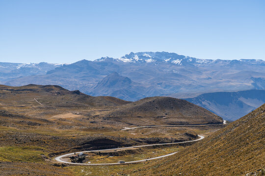 A winding asphalt road snakes through the high-altitude arid hills of the Peruvian Andes, looking toward the majestic Mismi mountain range under a clear blue sky