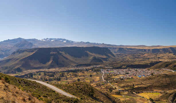 A panoramic view overlooks the town of Chivay nestled in the Colca Canyon valley, with the rugged Mismi mountain range rising in the background under a clear sky in Peru