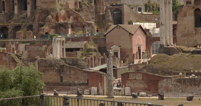 Detailed view of ancient ruins in the Roman Forum, showcasing fragmented stone columns, old brick walls, a small historic building, and the layered archaeological remains of the Roman Empire.