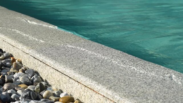 A close-up, selective focus shot of a luxury overflow swimming pool edge made of granite stone, with clear water running over it down into a drainage channel decorated with river pebbles.