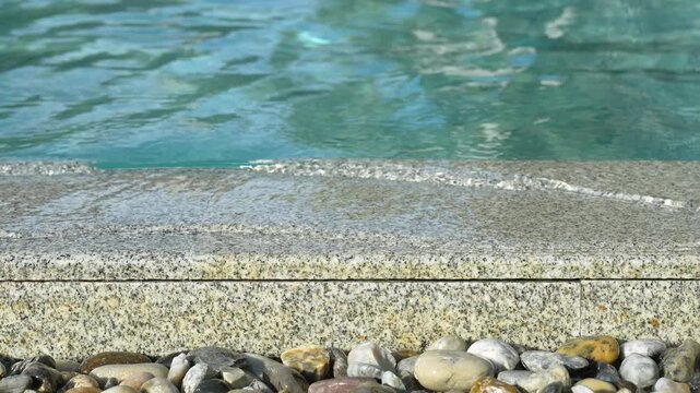 A close-up, selective focus shot of a luxury overflow swimming pool edge made of granite stone, with clear water running over it down into a drainage channel decorated with river pebbles.