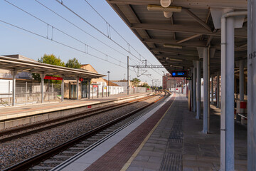 Obraz premium A suburban commuter rail station in Valencia, Spain, featuring covered platforms, overhead electric wires, digital departure boards, and passengers waiting under warm evening sunlight.