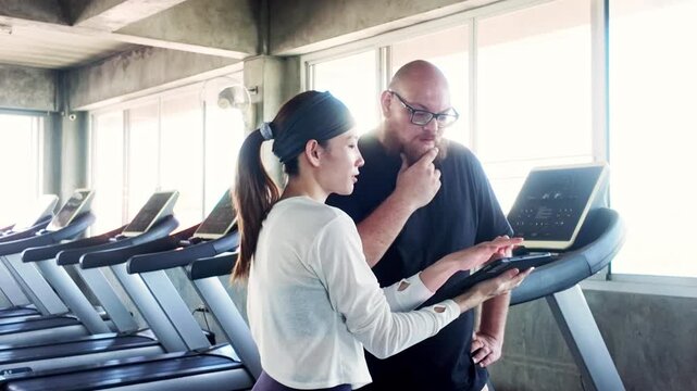 A fitness trainer shows a client treadmill data at a gym, planning a cardio workout beside a row of machines under bright windows.