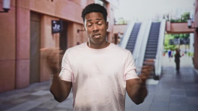 Man with hands raised shrugs in a building atrium by escalator with blurred pedestrians in view; uncertainty.