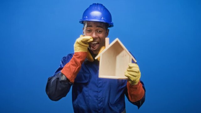 Man builder smiling and holding wooden house model while pointing with gloved hand in studio; pride homeownership.