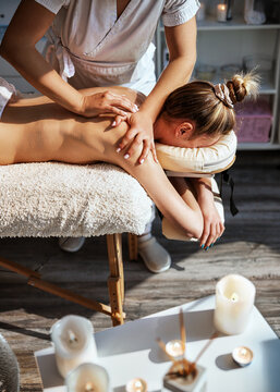 Manicured therapist, hands press into a womans shoulder area as she rests face down on a cream fleece covered table with lit candles and decorative items blurred softly in the foreground.
