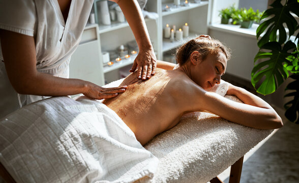 A therapist works a golden shimmer scrub across a relaxed womans bare back as she rests on a plush white towel covered table, surrounded by lit candles and monstera leaves in warm spa light.