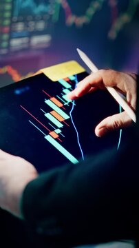 analyst hand holding tablet pointing digital pen dynamic candlestick chart displaying stock market price trends volatility. In the background, another screen with blurred multi colored financial graph