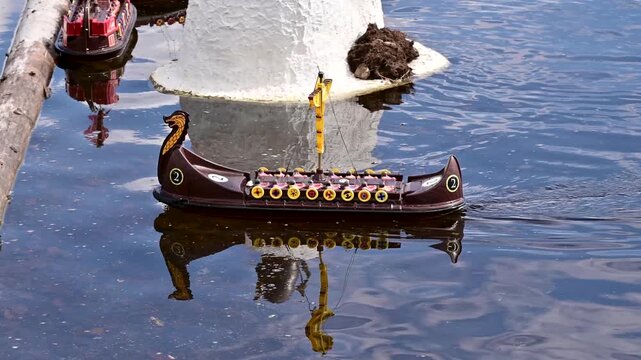 Toy viking longship remote controlled boat sailing on pond. Miniature brown plastic viking ship model floats across calm dark water surface. Slow motion 4K footage