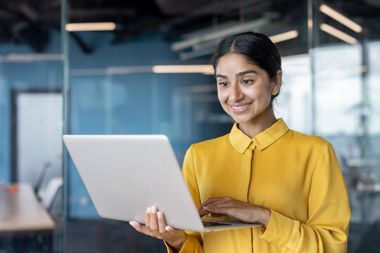 Close-up photo of a smiling young woman standing in the office, holding a laptop and typing