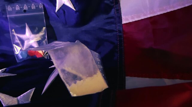 Close up shot of small plastic zip lock bags containing what appears to be narcotics like cocaine and ecstasy pills, lying on an American flag, symbolizing the drug crisis in the USA