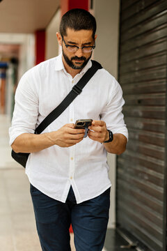 Middle aged man in smart casual outfit checking smartphone while walking along quiet urban street in Brazil with authentic lifestyle and natural light.