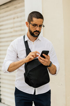 Middle aged man in smart casual outfit checking smartphone while walking along quiet urban street in Brazil with authentic lifestyle and natural light.