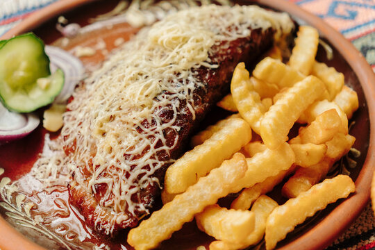 Traditional gourmet pljeskavica served with crispy French fries, grated cheese, fresh onion and cucumber. A hearty Balkan grilled meal presented on a rustic ceramic plate.