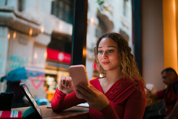 Fototapeta premium Smiling Caucasian woman browsing smartphone in cafe with tablet on table, mobile banking, online shopping, social media user, digital nomad lifestyle, 5G internet, authentic representation.