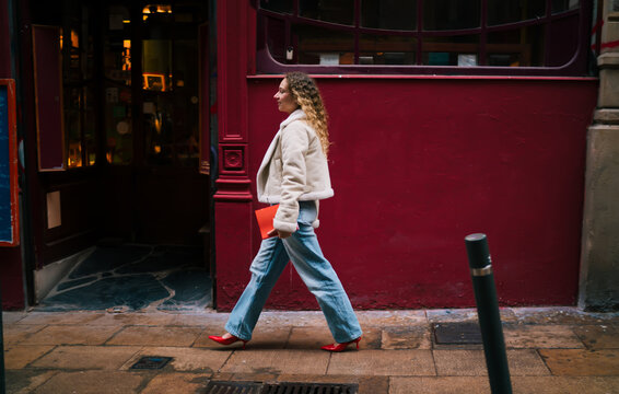 Full length profile of woman walking on city street past red facade, Caucasian model, white sherpa jacket, red shoes, blue jeans, urban lifestyle, authentic movement, sidewalk, architecture.