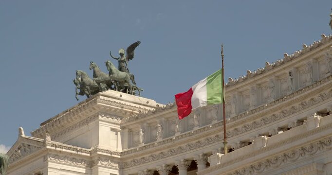 A close-up view of the Altare della Patria roof in Rome, Italy, showing the prominent Italian flag waving next to the majestic bronze quadriga statue against a clear, sunny blue sky.