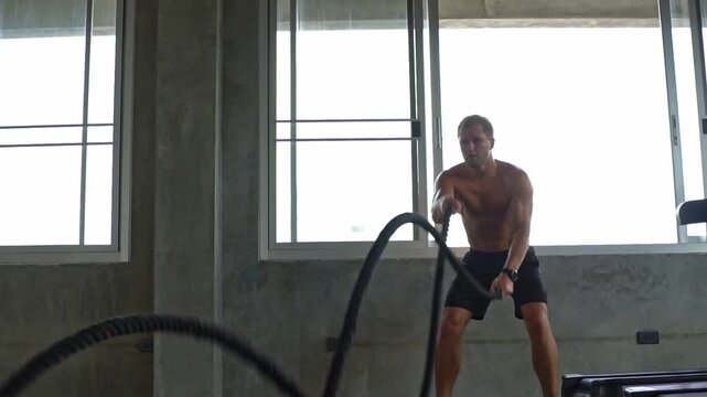 Shirtless muscular man exercising with battle rope at gym. Athletic male doing high intensity functional training fitness workout for strong body