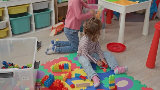 Girl kneels, hands in clear plastic toy box sorting colorful blocks on foam mat inside a playroom building; playful cooperation.