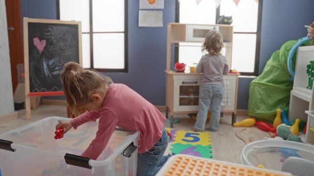Two girls play with plastic building blocks, reaching into a clear storage bin in a colorful playroom inside a building; playful concentration.