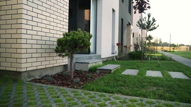 Modern townhouse exterior with a landscaped front yard featuring a small pine tree, lush green lawn, concrete slab walkway, and a permeable paver driveway for sustainable urban living