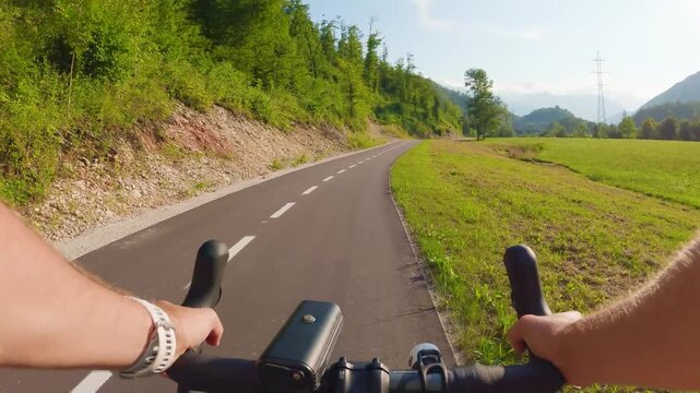 POV of cyclist riding on asphalt bike path in Slovenia. First person view of male hands on bicycle handlebar cycling on road near green alpine fields and mountains, active outdoor sport lifestyle