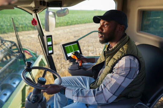 A farmer operates a combine harvester in a large field. The sky is clear, and crops grow well. He focuses on the controls while managing the machine.