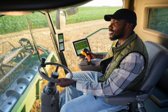 A farmer drives a large harvester in a field with green crops. The sun is shining as he uses a control panel to manage the machine.