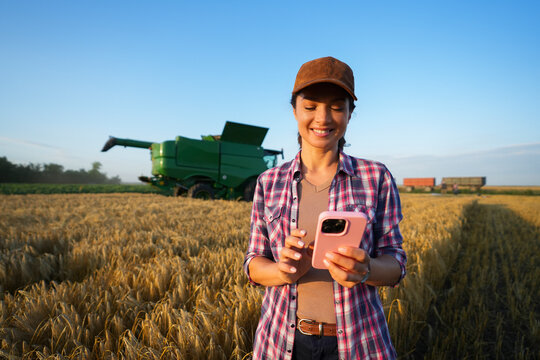 A farmer stands in a field of tall crops holding a smartphone. Behind her, a large green machine is visible. The sky is clear and blue with a hint of clouds.