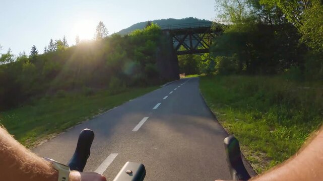 POV of cyclist riding on asphalt bike path under bridge in Slovenia. First person view of male hands on bicycle handlebar cycling on road near railway viaduct and fields, active sport lifestyle