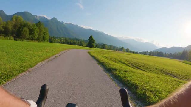 POV of cyclist riding on asphalt bike path in Slovenia. First person view of male hands on bicycle handlebar cycling on road near green alpine fields and mountains, active outdoor sport lifestyle