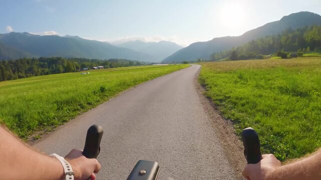 POV of cyclist riding on asphalt bike path in Slovenia. First person view of male hands on bicycle handlebar cycling on road near green alpine fields and mountains, active outdoor sport lifestyle