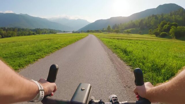 POV of cyclist riding on asphalt bike path in Slovenia. First person view of male hands on bicycle handlebar cycling on road near green alpine fields and mountains, active outdoor sport lifestyle