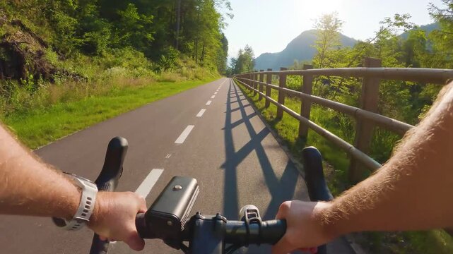 POV of cyclist riding on asphalt bike path at sunset in summer. Slovenia. First person view of male hands on bicycle handlebar cycling on road, wooden fence, river, green forest. Active sport outdoor
