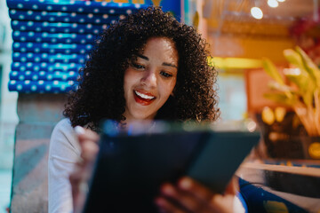 Cheerful young Hispanic woman with curly hair drawing on digital tablet screen with electronic stylus pencil, creative freelance and modern technology lifestyle concept, close up © BullRun